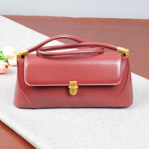 May include: A burgundy handbag with a top handle and a rectangular shape. The bag features a gold-coloured clasp and handle attachments. The bag is made of a smooth material and has a flap closure. The bag is sitting on a grey surface.