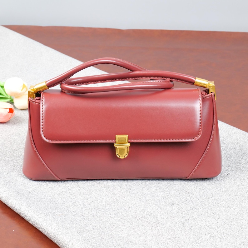 May include: A burgundy handbag with a top handle and a rectangular shape. The bag features a gold-coloured clasp and handle attachments. The bag is made of a smooth material and has a flap closure. The bag is sitting on a grey surface.