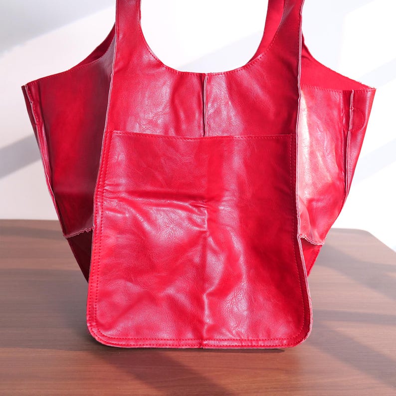 K&ouml;nnte beinhalten: Eine rote Ledertasche mit einer gro&szlig;en Vordertasche. Die Tasche hat zwei Henkel und eine rechteckige Form. Das Leder hat einen leichten Glanz. Die Tasche ist leer. Die Tasche steht auf einer braunen Oberfl&auml;che.