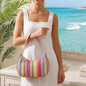 May include: A crescent-shaped handbag featuring a woven texture with vertical stripes in vibrant rainbow colors. The bag includes light pink leather-like trim, a matching shoulder strap, and a small circular gold-toned metal charm attached to the side.