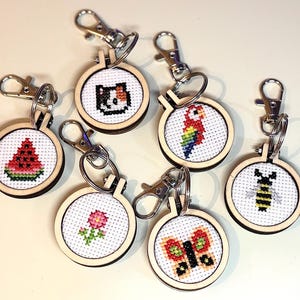 Puede incluir: Una colección de llaveros hechos a mano con punto de cruz. Cada llavero presenta un diseño de punto de cruz en miniatura dentro de un aro de madera. Los diseños incluyen una sandía, un gato, un loro, una flor, una mariposa y una abeja.