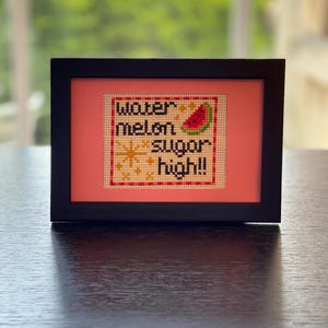 May include: A framed cross-stitch embroidery featuring the text "watermelon sugar high!!" in black thread. The design includes a small watermelon slice, yellow starburst accents, and a red and white checkered border on a cream fabric background.