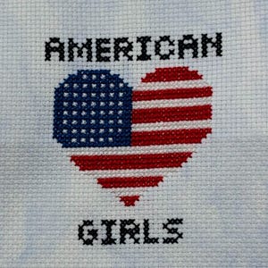 May include: Cross-stitch design showing a heart shaped like the American flag. The words "AMERICAN" and "GIRLS" are stitched in black above and below the heart. The background is a light blue fabric.