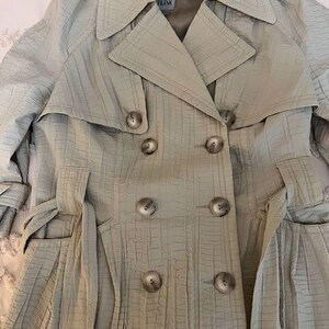 May include: A light beige trench coat with a textured, grid-like pattern. The double-breasted coat features large buttons, a notched collar, and a belt. The label reads "PEOPLE LIKE FRANK".