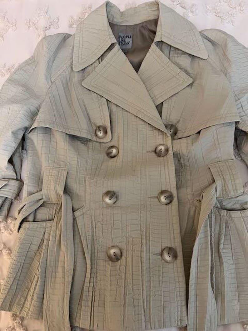May include: A light beige trench coat with a textured, grid-like pattern. The double-breasted coat features large buttons, a notched collar, and a belt. The label reads "PEOPLE LIKE FRANK".