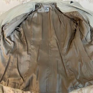 May include: Open view of a light green jacket with a textured exterior and a silky, tan interior lining. The jacket has a classic design with a collar and a belt. The label reads "People by Frank".