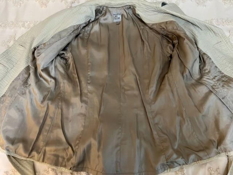 May include: Open view of a light green jacket with a textured exterior and a silky, tan interior lining. The jacket has a classic design with a collar and a belt. The label reads "People by Frank".
