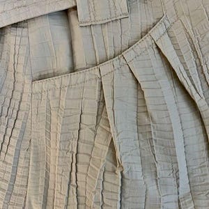 May include: Close-up of a beige jacket with a textured, pleated design. The jacket features a tailored fit with a belt and a large button. The fabric has a subtle sheen, and the stitching is visible.