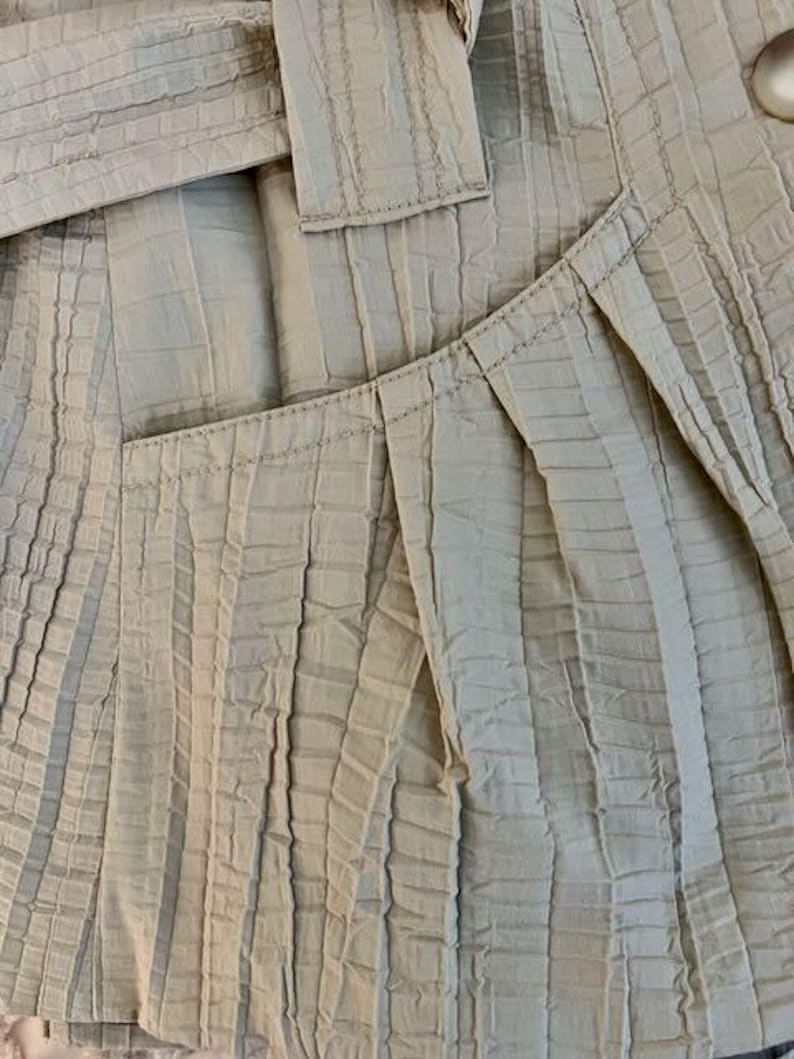 May include: Close-up of a beige jacket with a textured, pleated design. The jacket features a tailored fit with a belt and a large button. The fabric has a subtle sheen, and the stitching is visible.