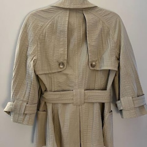 May include: A beige trench coat with a textured pattern, hanging on a metal hanger. The coat features a belt, two front pockets, and button details. The sleeves are three-quarter length.