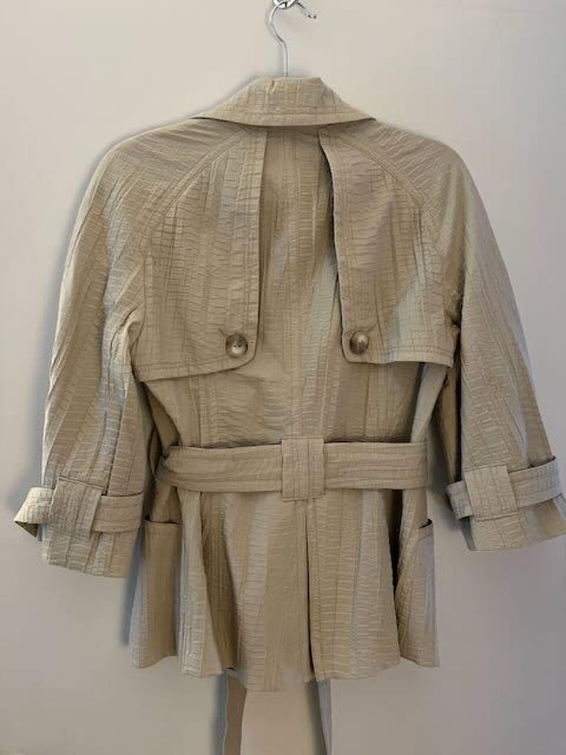 May include: A beige trench coat with a textured pattern, hanging on a metal hanger. The coat features a belt, two front pockets, and button details. The sleeves are three-quarter length.