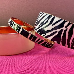 May include: Three bangle bracelets with gold-tone rims. One is a wide solid cream-colored bangle, one is a thin bangle with a black and white zebra print, and one is a wide bangle featuring a bold black and white zebra stripe pattern throughout.