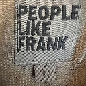 May include: Close-up of a gray fabric label with the words "PEOPLE LIKE FRANK" in dark gray block letters. Below the text is a smaller label with the letter "L". The background is a tan fabric.