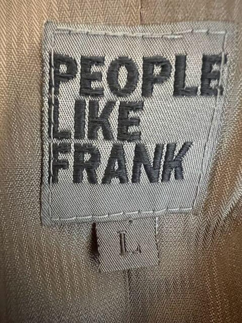 May include: Close-up of a gray fabric label with the words "PEOPLE LIKE FRANK" in dark gray block letters. Below the text is a smaller label with the letter "L". The background is a tan fabric.