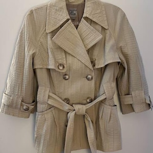 May include: A beige trench coat with a double-breasted design, featuring a notched collar, button closures, and a matching belt tied at the waist. The coat has three-quarter sleeves and is hanging on a metal hanger.