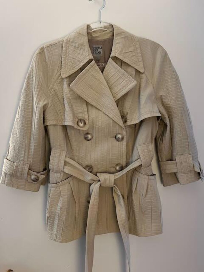 May include: A beige trench coat with a double-breasted design, featuring a notched collar, button closures, and a matching belt tied at the waist. The coat has three-quarter sleeves and is hanging on a metal hanger.