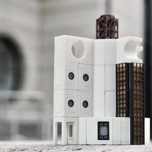 Può includere: Un modello architettonico bianco con una struttura a traliccio marrone scuro sulla parte superiore e una sezione a griglia nera e oro. Il modello presenta aperture circolari e quadrate e piccoli dettagli rotondi neri. L'edificio è su una superficie chiara.