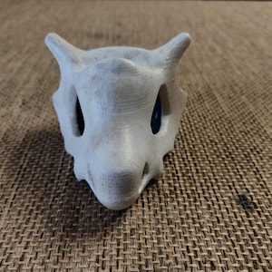 May include: A light grey, stylized skull with large eye sockets and two pointed horns. The skull has a textured surface and dark blue accents in the eye sockets. The object is likely a decorative item or part of a costume.