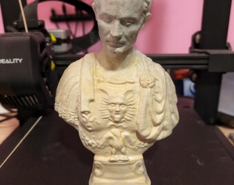 May include: A light beige bust of a man with short, curly hair. The bust features detailed carvings of a face, chest, and shoulders. The chest has a detailed design of a face and an eagle. The bust is on a black surface.