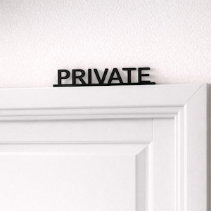 May include: A black "PRIVATE" sign is mounted above a white door. The sign is in a sans-serif font. The door has a classic, paneled design with a white finish, and is set against a white wall.