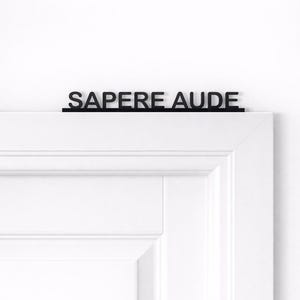 Könnte beinhalten: Ein schwarzes Metallschild mit der lateinischen Phrase "SAPERE AUDE", das über einem weißen Türrahmen angebracht ist. Die Buchstaben des Schildes sind in einer einfachen, serifenlosen Schrift gehalten, was eine minimalistische Ästhetik erzeugt. Der Türrahmen ist weiß, mit einem klassischen Paneeldesign.