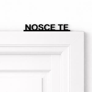 Può includere: Un cartello nero con le parole "NOSCE TE" in un font sans-serif è montato sopra un telaio di porta bianco. Il cartello è un elemento decorativo, probabilmente per l'interior design.
