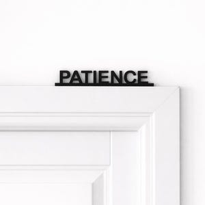 Könnte beinhalten: Ein schwarzes Holzschild mit der Aufschrift "PATIENCE" befindet sich auf einer weißen Tür. Der Text ist in Großbuchstaben geschrieben. Die Tür ist weiß mit einem eingelassenen Paneeldesign. Nahaufnahme.