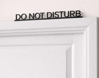 Puede incluir: Letrero negro de "DO NOT DISTURB" sobre una puerta blanca. El diseño minimalista del letrero es ideal para un dormitorio u oficina. La puerta tiene un diseño de panel clásico.