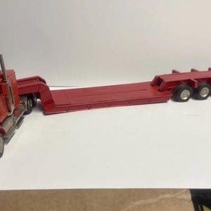 Puede incluir: Un camión y remolque de juguete rojo. El camión tiene una parrilla delantera detallada y una cabina con techo. El remolque es un diseño de plataforma con múltiples ejes. El juguete probablemente está hecho de metal fundido a presión.