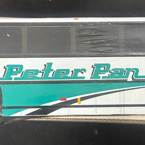 May include: A white and teal Peter Pan Trailways bus model. The bus has black windows, white wheels, and the American flag on the side. The text "Peter Pan Trailways" is displayed in teal and yellow.