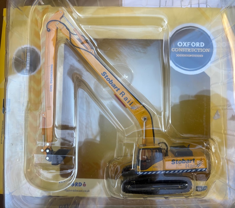 May include: A yellow and black model excavator toy with the text "Stobart Rail" and "Civil Engineering". The toy is packaged in a clear plastic container with the "Oxford Construction" logo.