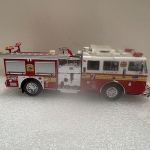 Könnte beinhalten: Ein rot-weißes Feuerwehrauto-Modell mit dem F.D.N.Y.-Logo, der Nummer 7 an der Seite, silbernen Metalldetails, einer auf dem Dach montierten Leiter, Blaulichtern und verschiedenen kleinen Aufklebern an den Seitenwänden des Fahrzeugs.