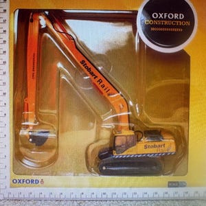 May include: A toy excavator model in a clear plastic package. The excavator is primarily orange and yellow, with black treads and the text "Stobart Rail" and "Oxford Construction". The model is labeled "Civil Engineering".