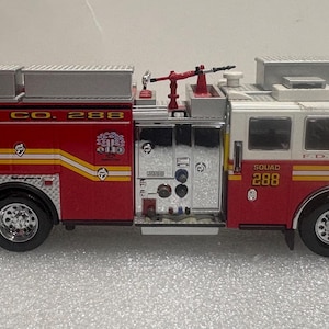 Op de afbeelding: Een rood en wit FDNY Squad Co. 288 brandweerwagenmodel. Het model heeft de tekst "FIRE" en "SQUAD CO. 288" in geel en wit. De truck heeft zilveren accenten, een ladder en zwaailichten.