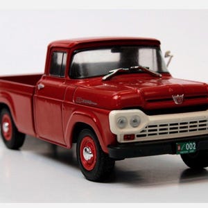 May include: A red vintage pickup truck model with a white grille and headlights. The truck has black tyres and a license plate. The model is a classic car collectible.