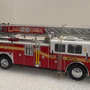 Puede incluir: Modelo de camión de bomberos rojo y blanco con una escalera. El modelo tiene el texto "LAG26DER" y "FDNY Fire Factory Harlem N.Y.". El camión tiene rayas amarillas y el número "36" en el lateral.