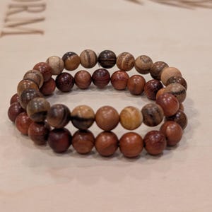 Puede incluir: Dos pulseras de cuentas de madera. Las cuentas son de varios tonos de marrón, desde el beige claro hasta el marrón rojizo oscuro. Las pulseras están hechas de cuentas redondas, ensartadas para formar una banda flexible. Las pulseras se exhiben sobre una superficie clara.