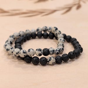 May include: Two beaded bracelets. One bracelet features black lava beads. The other bracelet has white beads with black spots. The bracelets are stacked on top of each other.