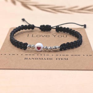 May include: Black macrame bracelet with a white bead featuring a red heart, silver beads, and a silver tube. The bracelet is on a brown card that says "I Love You" and "HANDMADE ITEM".