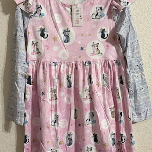 May include: A pink dress with a cat pattern, featuring long sleeves and ruffled shoulder details. The dress has a gathered skirt and is adorned with various cat illustrations, stars, and circles. The sleeves are light blue with a star pattern.