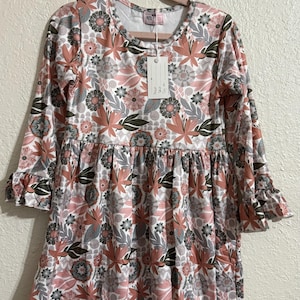 May include: A long-sleeved dress with a floral pattern in shades of pink, gray, and green on a white background. The dress has a round neckline, a gathered waist, and ruffled sleeves. The dress is hanging on a pink hanger.