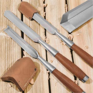 May include: A set of woodworking chisels with stainless steel blades and wooden handles. The chisels are arranged on a wooden surface, with leather blade protectors. The tools are designed for carving and shaping wood.