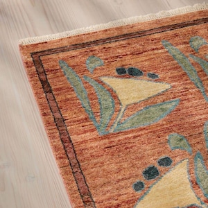 May include: Close-up of a terracotta-colored rug with a geometric floral pattern. The rug features a border with dark brown and red accents. The design includes light yellow, teal, and dark blue shapes. The rug has a fringed edge.