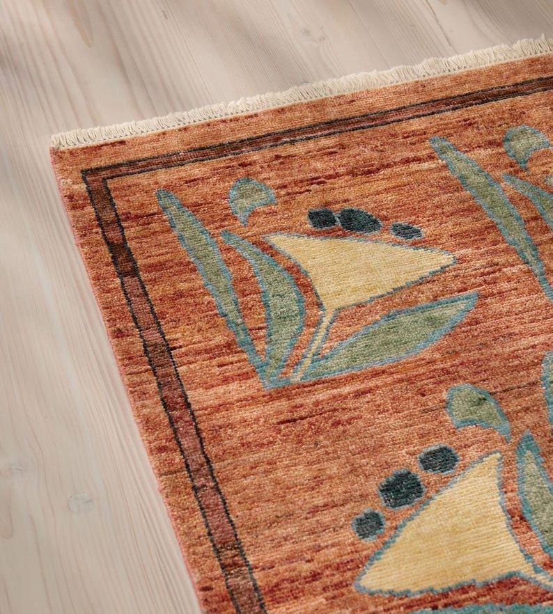 May include: Close-up of a terracotta-colored rug with a geometric floral pattern. The rug features a border with dark brown and red accents. The design includes light yellow, teal, and dark blue shapes. The rug has a fringed edge.