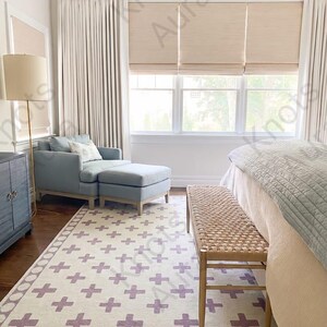 Puede incluir: Un interior de dormitorio con un sillón azul claro con su reposapiés a juego, un banco trenzado y una alfombra color crema con un patrón repetido de cruces moradas. La habitación incluye cortinas color crema y estores romanos beige.