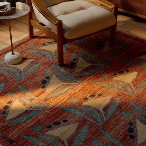 May include: A rectangular area rug with a floral pattern in shades of orange, beige, and teal. The rug is placed on a wooden floor, with a wooden chair and side table in the background. The rug's design features stylized flowers.