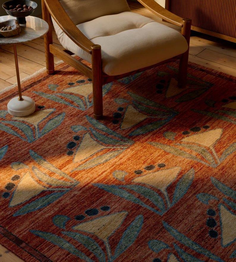 May include: A rectangular area rug with a floral pattern in shades of orange, beige, and teal. The rug is placed on a wooden floor, with a wooden chair and side table in the background. The rug's design features stylized flowers.