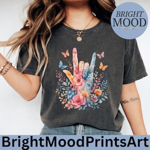 Può includere: T-shirt grigio scuro con un design colorato del segno della mano, adornato con fiori e farfalle. Il testo "BrightMoodPrintsArt" è in basso, con un logo circolare con lo stesso testo in alto a destra.
