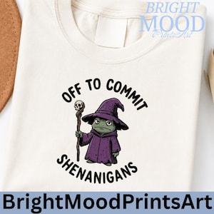 May include: Cream-colored t-shirt with a graphic of a grumpy wizard frog holding a staff. The text reads "OFF TO COMMIT SHENANIGANS" in black. The wizard wears a purple robe and hat. The brand name "Bright Mood Prints Art" is at the top and bottom.
