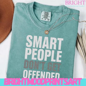 Op de afbeelding: Een turquoise Comfort Colors T-shirt met de tekst "SMART PEOPLE DON'T GET OFFENDED" in witte en grijze letters. Een zilveren ketting met een kleine hanger ligt op het shirt. Het shirt is maat small.